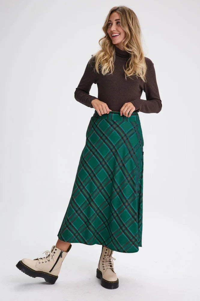 A woman wearing the Montebello Skirt by Message Factory in Green, a plaid maxi skirt featuring a flared shape, elastic at the back of the waist, eco friendly fabric and a bias cut - plus pockets. She's wearing it with the Gatineau Turtleneck in Brown and standing in front of a white background.  