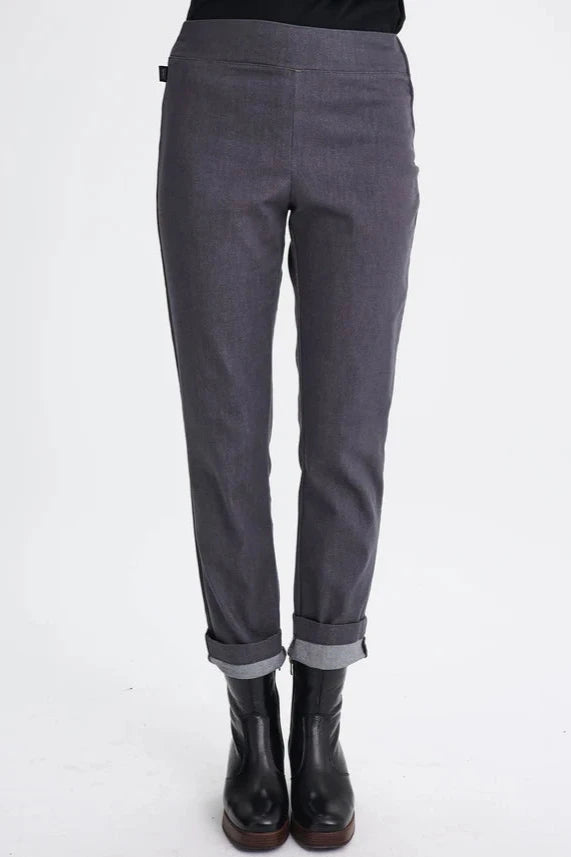 Waist-down view of a woman wearing the Potton Pants by Message Factory in Gray Denim,  slim cut pants in stretchy denim with a wide waistband and a side zipper, made from recycled cotton and polyester. She's standing in front of a white background. 