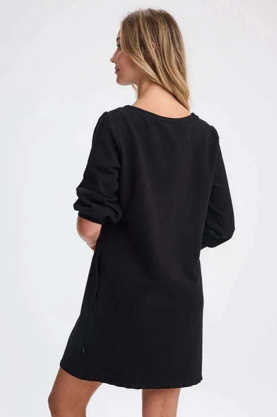 Back view of a woman wearing the Stukely Dress by Message Factory in Black, a semi-fitted bamboo/organic cotton fleece dress with a boat neck, 3/4 sleeves, side pockets, and a mid-thigh length. She is standing in front of a beige background. 