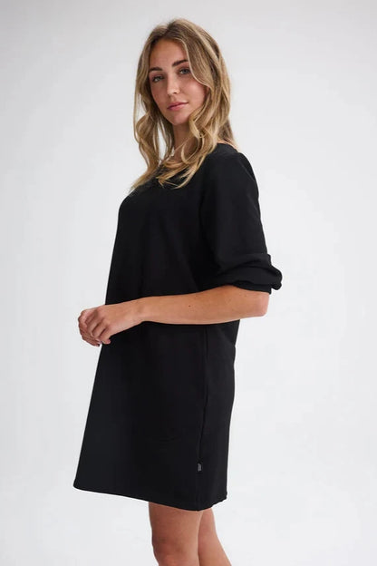 Side view of a woman wearing the Stukely Dress by Message Factory in Black, a semi-fitted bamboo/organic cotton fleece dress with a boat neck, 3/4 sleeves, side pockets, and a mid-thigh length. She is standing in front of a white background. 