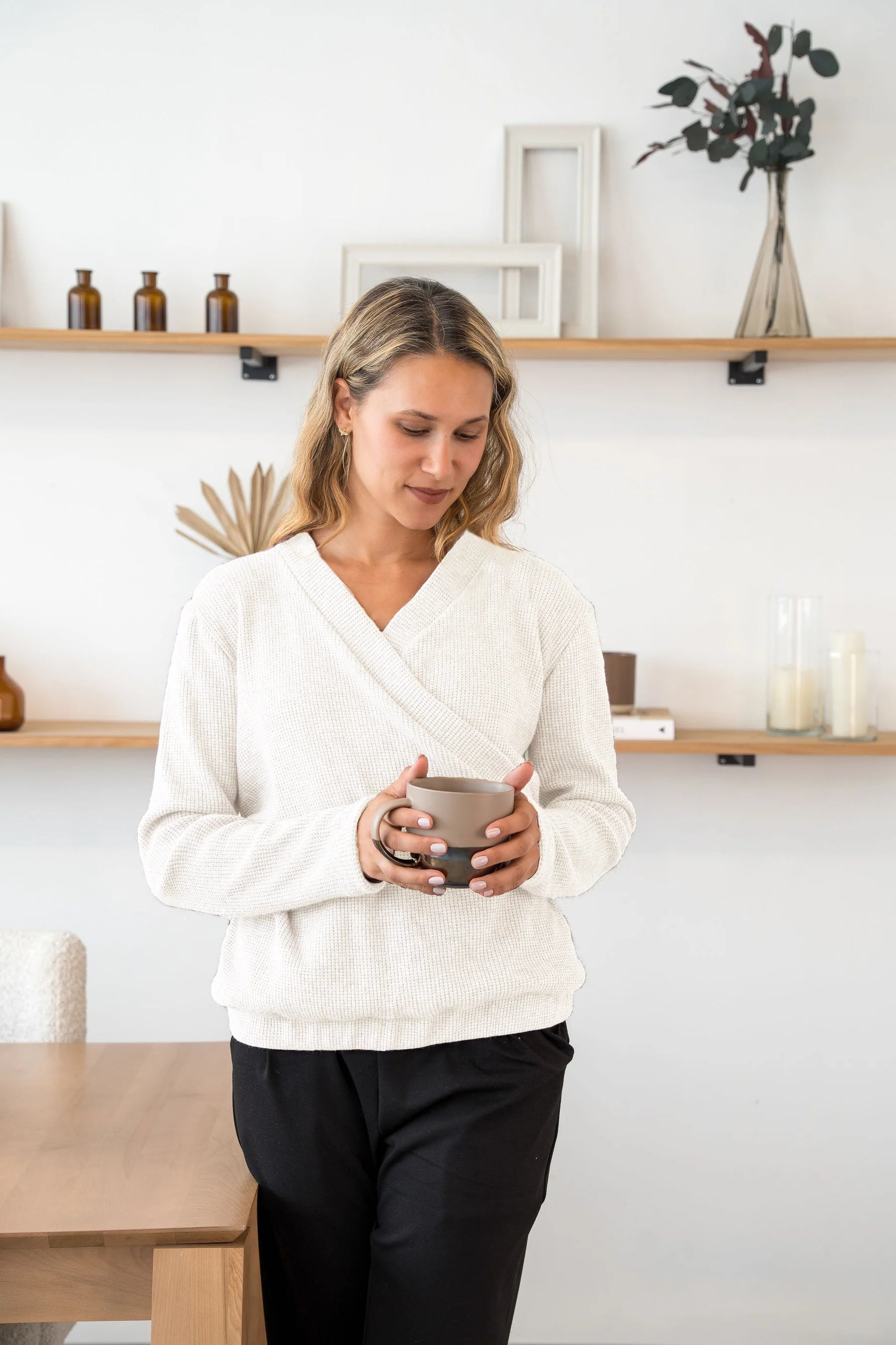 A woman wearing the Mimosa Sweater by Cherry Bobin in Ivory, a chunky knit sweater with a faux-wrap neckline, long slightly puffed sleeves, and an elastic waistband. She is wearing it with black pants and holding a mug while standing by a table. 