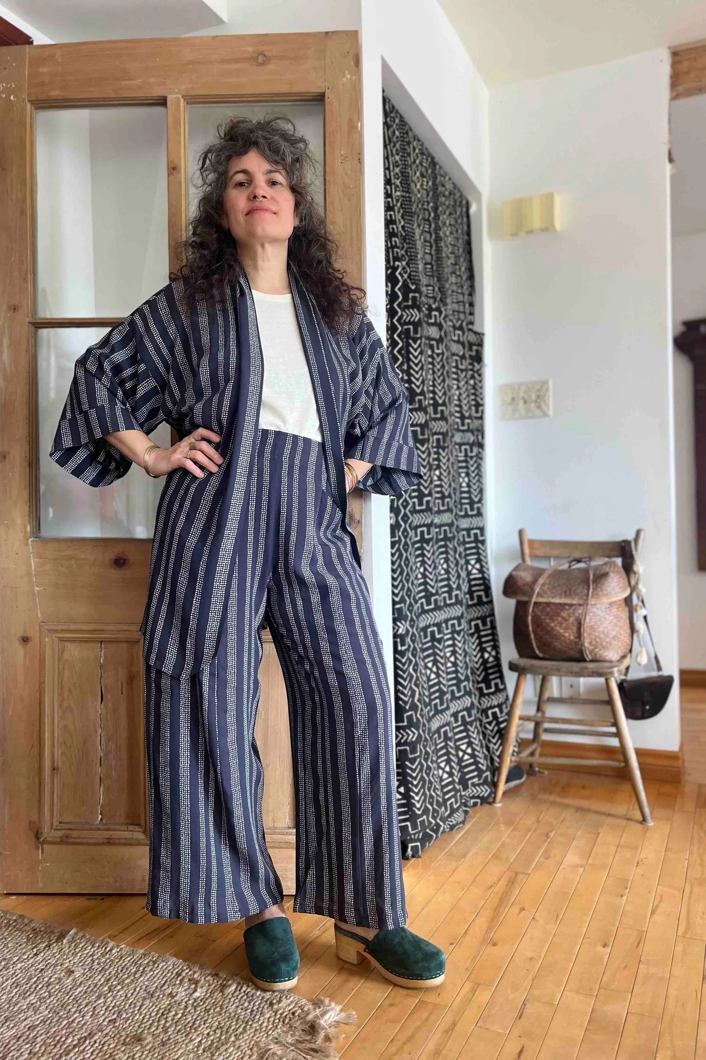 A woman wearing the Mirano Jacket by Kazak in Charcoal Dots print, an open Kimono-inspired jacket that falls to mid-thigh and has wide 3/4 sleeves. She is wearing it with white top top and matching pants and is standing in front of a wooden door.
