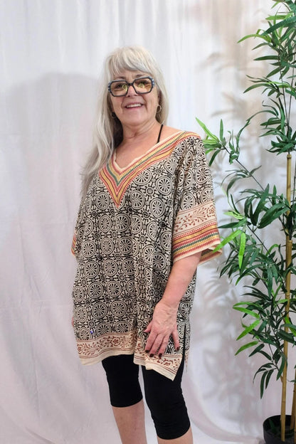 A woman wearing the Boho Tunic Dress (V-neck) by MOOVment in Black floral, a loose fitting piece with rainbow 
 coloured embroidery around the V-neck and wide short sleeves. She is standing in front of a white curtain. 
