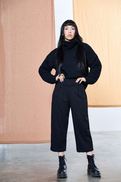 A woman wearing the Nathan Pants by Melow in Black speckled fabric, 7/8 length wide legged pants with a high pull-on waist band and front pleats. She is wearing them with a black turtleneck sweater and standing in front of a pastel background. 