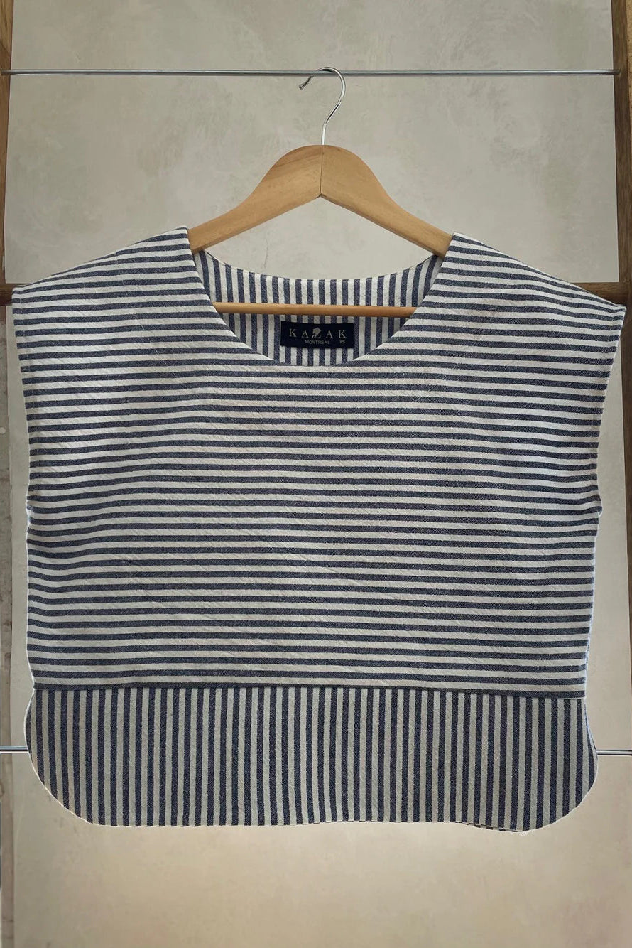 The Tulipe Top by Kazak in Black, a short top with a round neck, short extended sleeves, horizontal stripes across the chest and a band of vertical stripes across the bottom, with a rounded hem and side slits, is shown hanging from a wooden hanger against a white wall. 