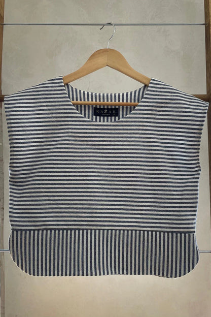 The Tulipe Top by Kazak in Black, a short top with a round neck, short extended sleeves, horizontal stripes across the chest and a band of vertical stripes across the bottom, with a rounded hem and side slits, is shown hanging from a wooden hanger against a white wall. 