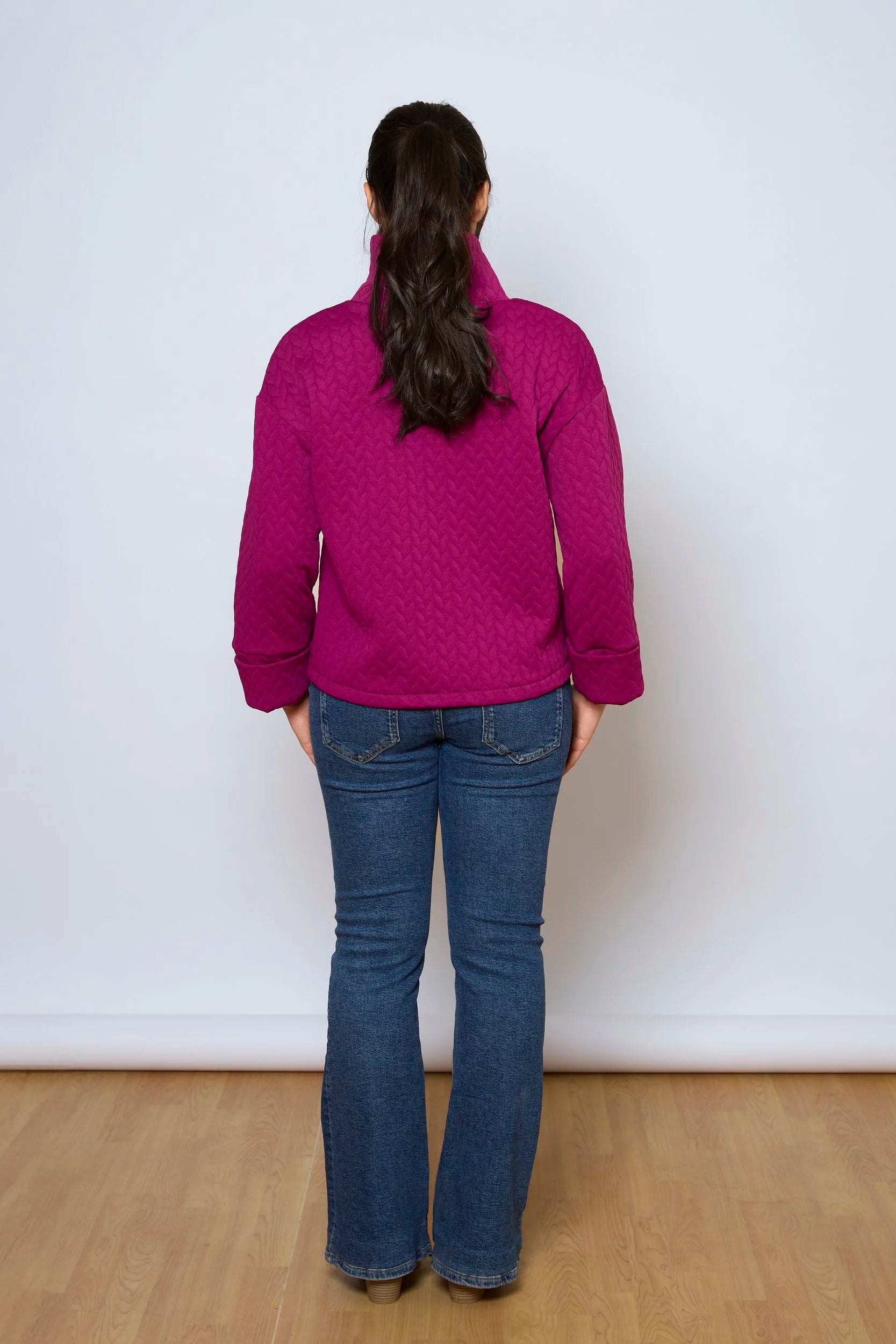 Back view of a woman wearing the Nori Top by Tangente in Magenta, a relaxed top in quilted fabric with a large cowl neck and cuffed drop sleeves. She is wearing it with jeans and standing in front of a white background. 