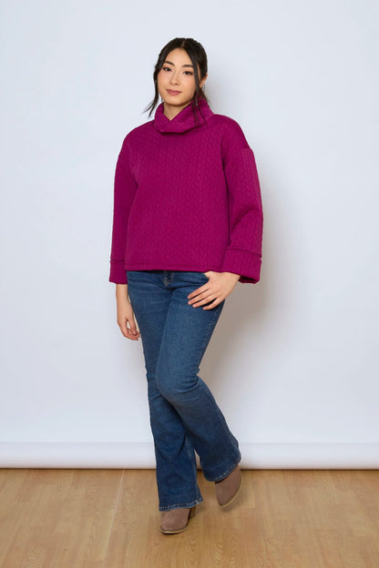 A woman wearing the Nori Top by Tangente in Magenta, a relaxed top in quilted fabric with a large cowl neck and cuffed drop sleeves. She is wearing it with jeans and standing in front of a white background. 