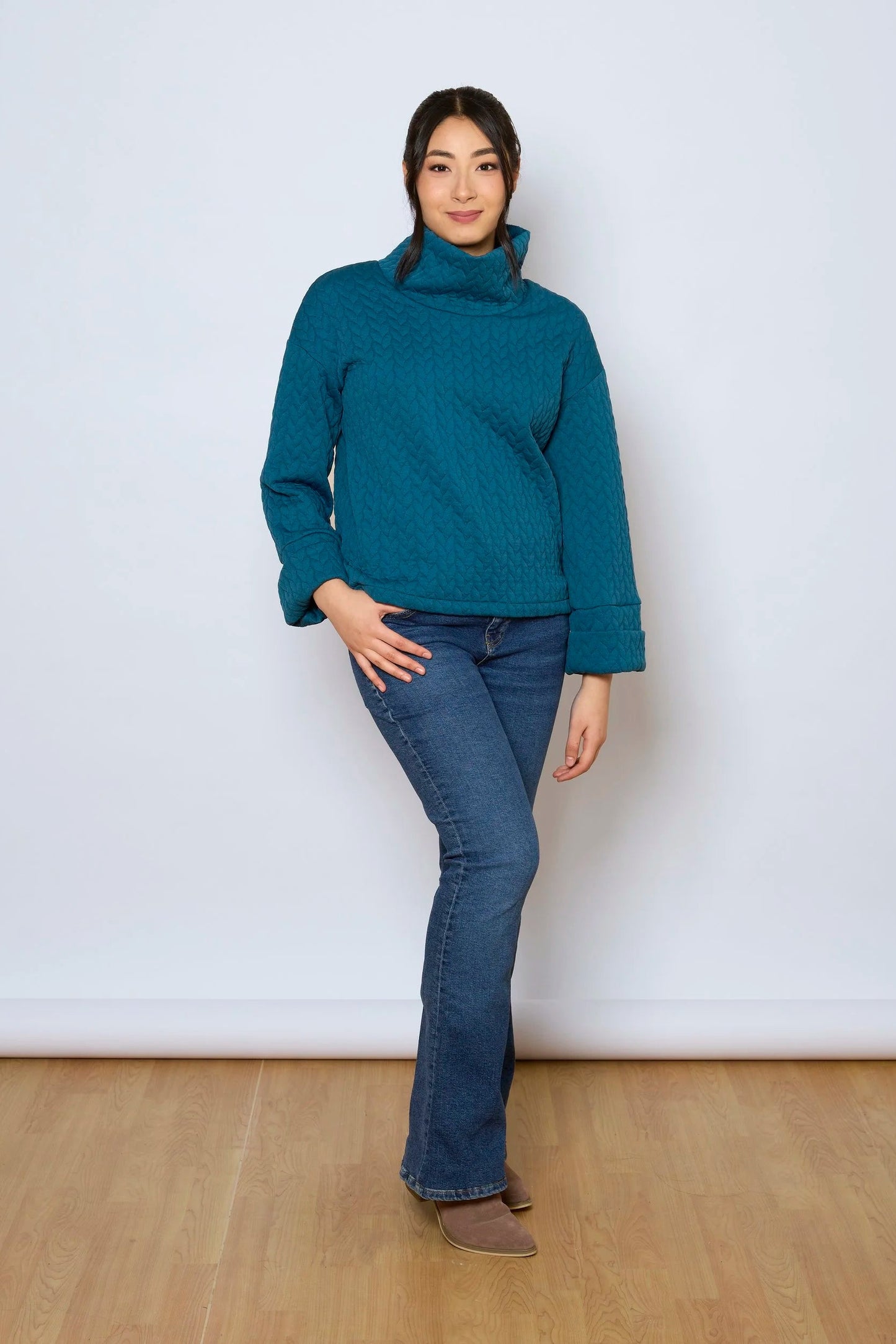 A woman wearing the Nori Top by Tangente in Peacock, a relaxed top in quilted fabric with a large cowl neck and cuffed drop sleeves. She is wearing it with jeans and standing in front of a white background. 