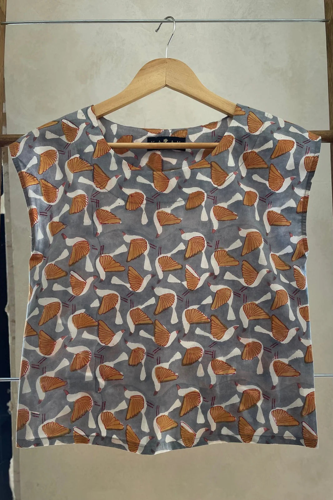 The Sandrigo Top from Kazak in Birds  print, a cotton top with short extended sleeves and a wide round neck, is shown on a wooden hanger against a grey background. 
