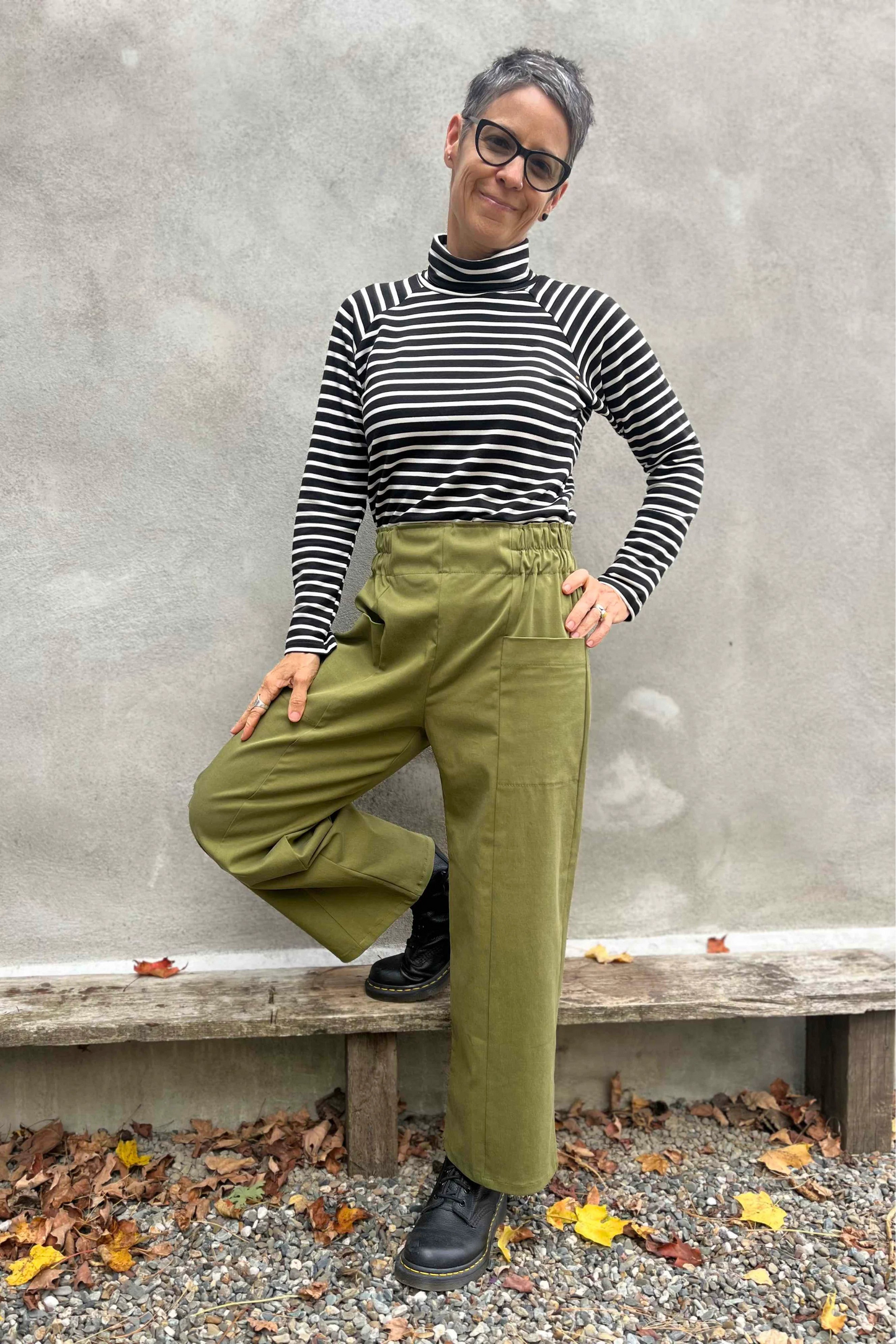 A woman wearing the Olea Pants by 
Kazak in Olive, wide-legged ankle-length pants with a flat-front elastic waist and large patch pockets. She is wearing them with the Oris Top in Blue Stripe and standing outside by a grey wall. 