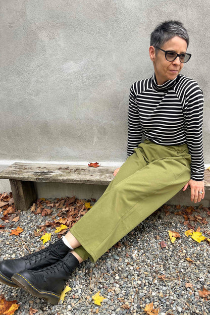 A woman wearing the Olea Pants by 
Kazak in Olive, wide-legged ankle-length pants with a flat-front elastic waist and large patch pockets. She is wearing them with the Oris Top in Blue Stripe and sitting outside by a grey wall. 