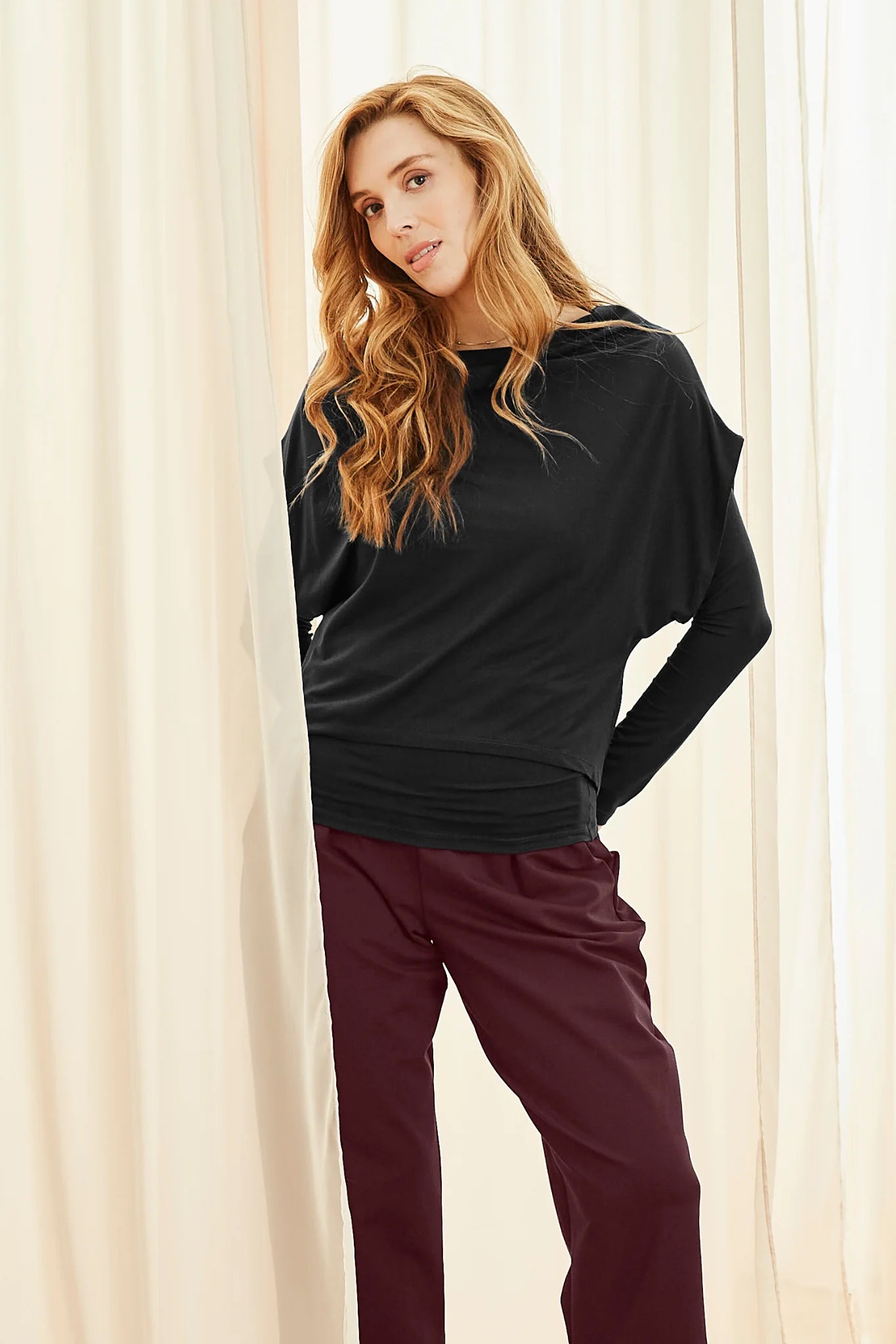 A woman wearing the Pacifique Top by Melow in Black, a two-layered top with a fitted and long-sleeved under-layer and a short-sleeved and loose-fitting over-layer. She is wearing it with burgundy pants and standing in front of white curtains. 