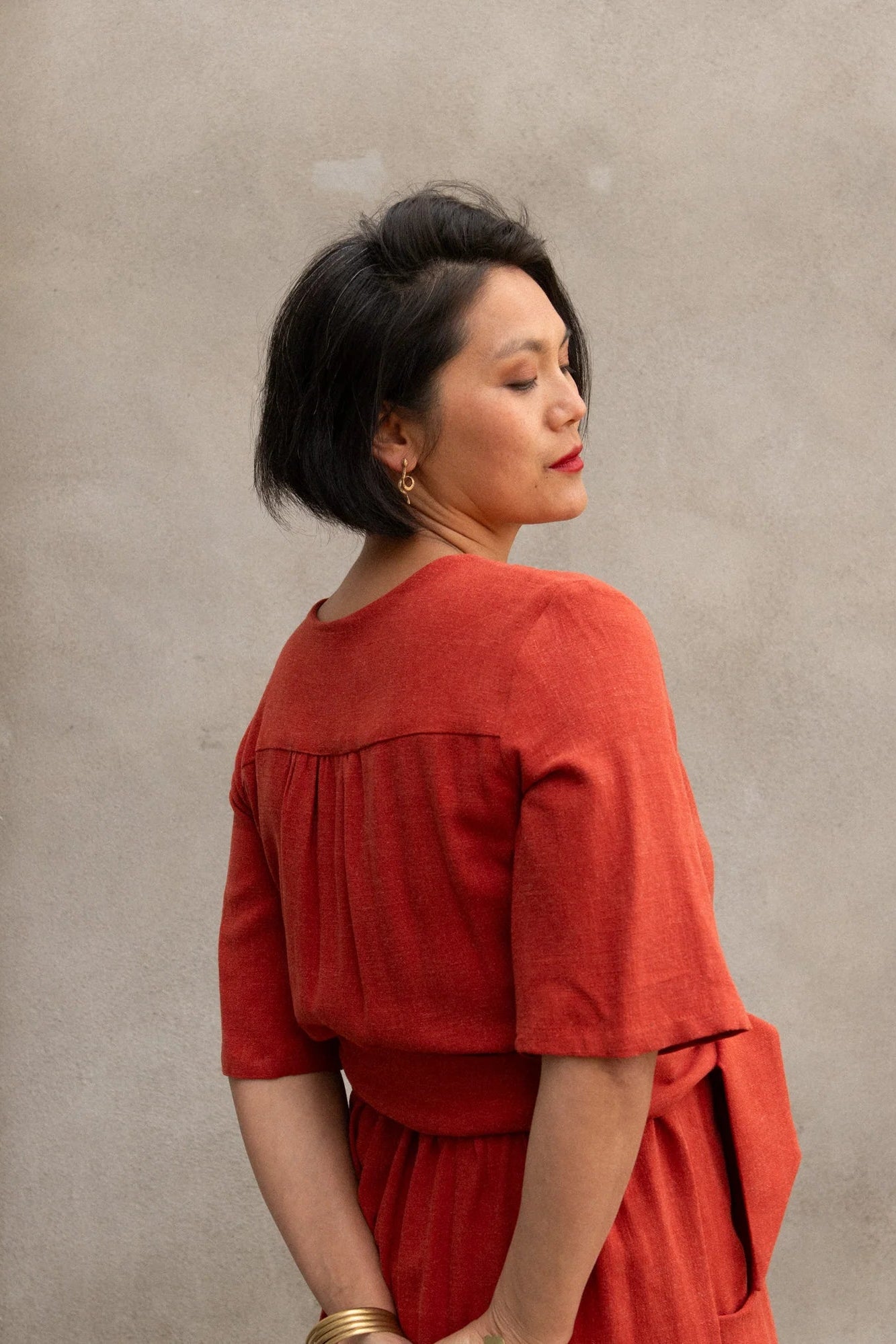 Back view of a woman wearing the Helleborus wrap Dress by Kazak in Paprika, standing outside by a white curtain and a cement wall. 