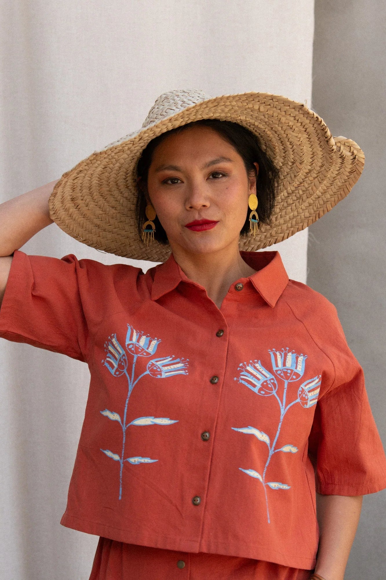 Waist up view of a woman wearing the Hosta Screen-printed Blouse by Kazak in Red with Pysanka, with hand-screen-printed flowers, mid-length raglan sleeves, a classic collar, and a button front. She is wearing it with a matching skirt and straw hat and standing outside by a grey wall. 