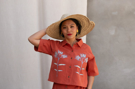 Waist up view of a woman wearing the Hosta Screen-printed Blouse by Kazak in Red with Pysanka, with hand-screen-printed flowers, mid-length raglan sleeves, a classic collar, and a button front. She is wearing it with a matching skirt and straw hat and standing outside by a grey wall. 