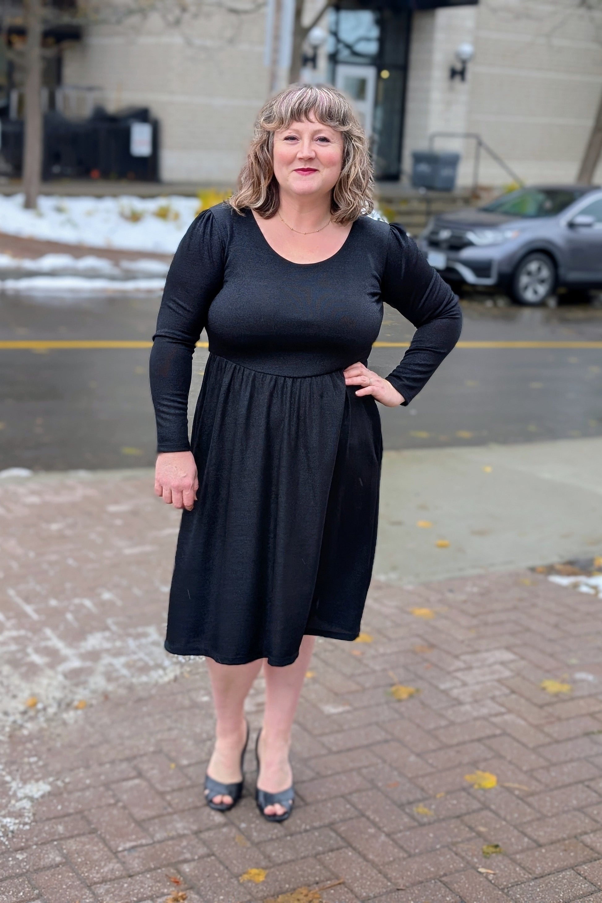 Woman wearing the Eve Lavoie 
Kennedy dress in Sparkly Black standing on a sidewalk with a building and car in the background.