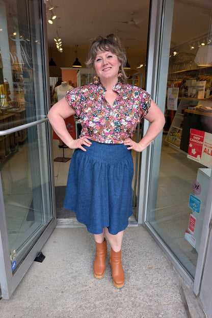 A woman wearing the Melow Margot Denim Skirt, a knee-length skirt with a wide pointed waistband, A-line shape, and invisible side zipper. She is wearing it with the Dotty Petal Top and standing in the doorway at Flock. 