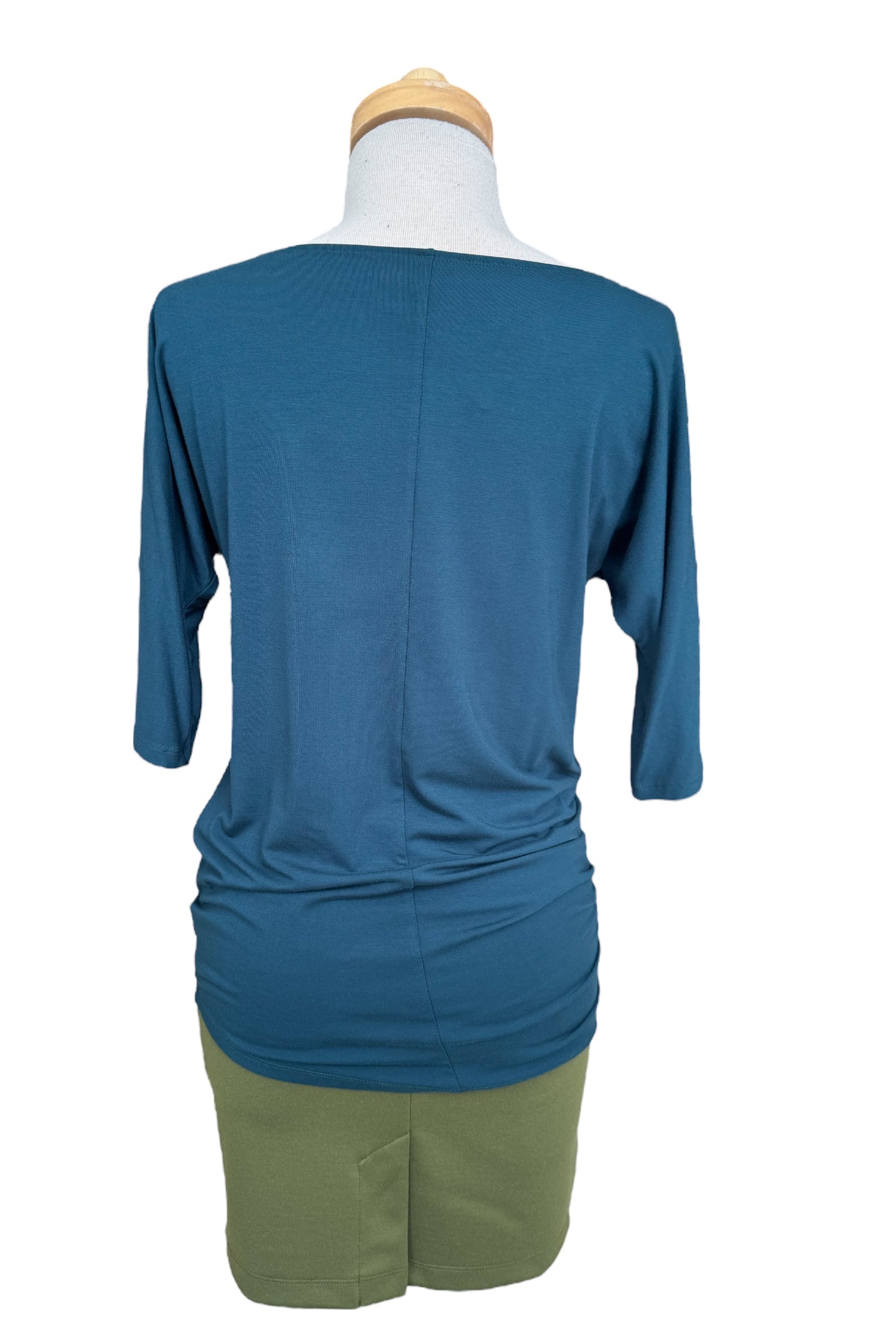 A back view of the Bettina Top by MOOVment in Teat, with boat neck, 3/4 sleeves, and gathered sides, is shown with a green skirt on a mannequin in front of a white background. 