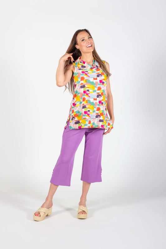 A woman wearing the Cailey Tunic by Pure Essence in a geometric Citrus/Coral pattern with purple pants, standing in front of a white back ground