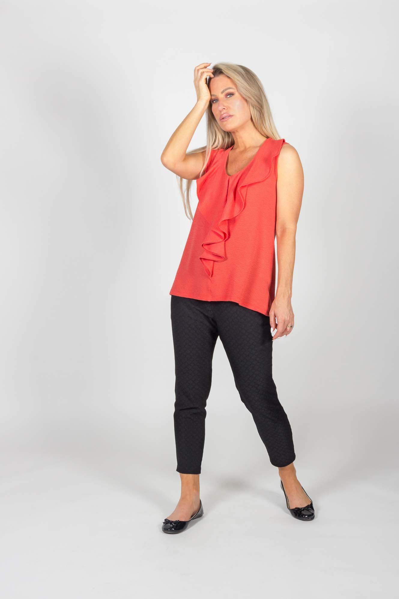 A woman wearing the Carson Top by Pure Essence in Coral, with the Celeste Pants in Black, standing in front of a white background 