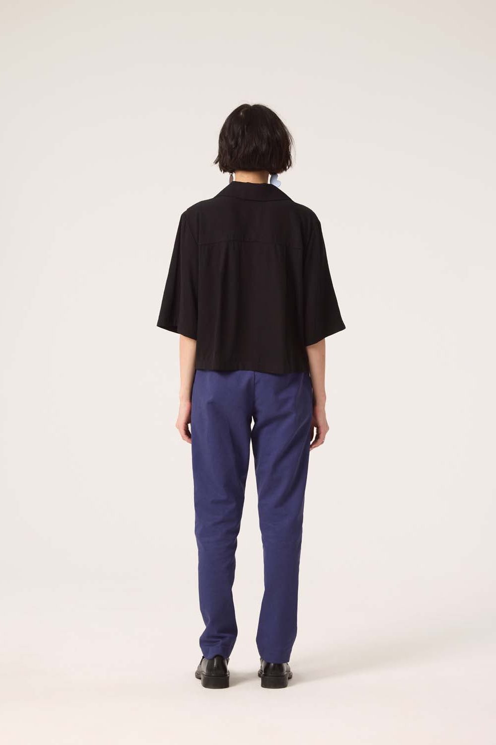 Back view of a woman wearing the Ritual Top by Cokluch in Black with blue pants, standing in front of a white background 