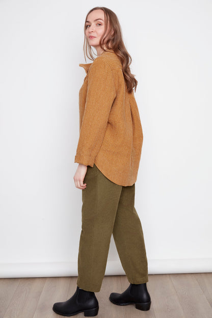 Side view of a woman wearing the Charlie Top by Julie in Ginger Stripes, with an asymmetrical button placket at the neckline and hi-low hemline with side slits. She's wearing them with green pants and standing in front of a white background. 