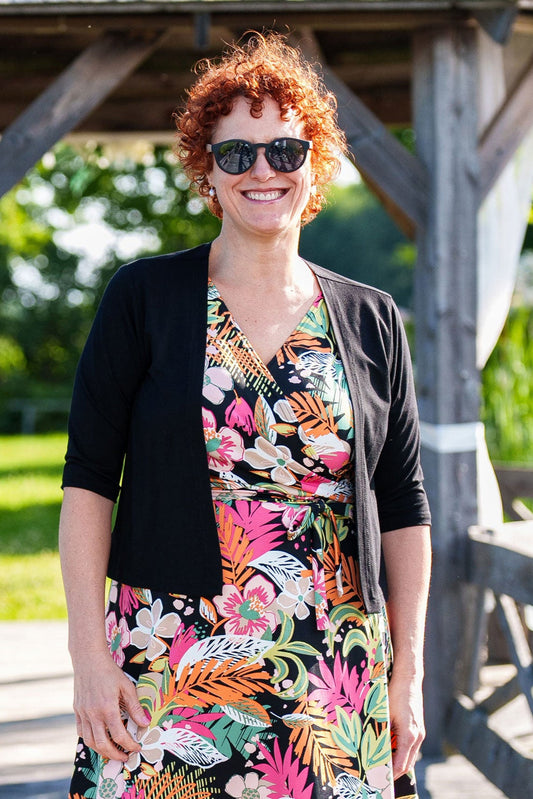3/4 view of a woman wearing the Salvador Bolero in Black from Rien ne se Perd, featuring 3/4 sleeves and an angled hem, over the Macarena Dress. She is standing on a dock in front of a wooden gazebo. 