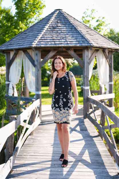 A woman wearing the Classic Dress by Rien ne se Perd in Black and Gold, a semi-fitted black  sheath dress with a gold floral pattern, short sleeves, and an above the knee length. She is standing on a wooden bridge in front of a pagoda. 