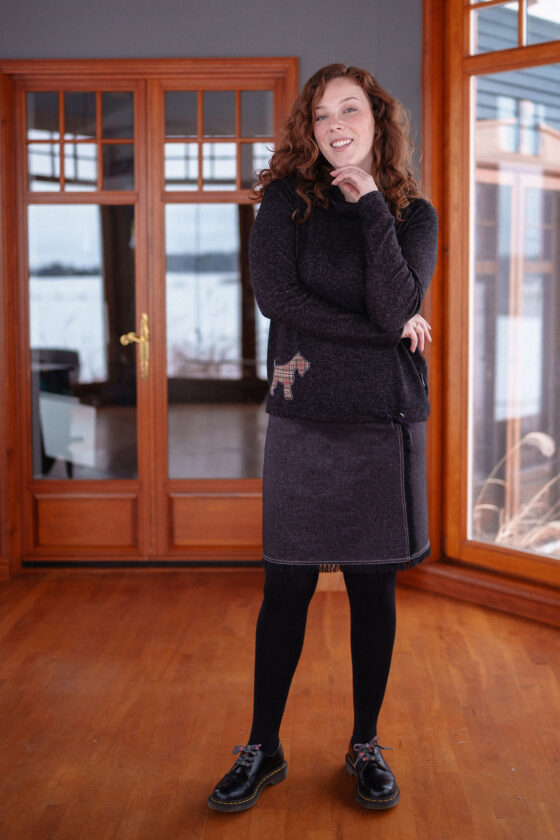 A woman wearing the Gustave Sweater in Charcoal with a plaid Scottish Terrier applique, and a denim skirt, standing by a picture window. 