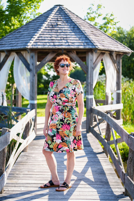 A woman wearing the Macarena Dress by Rien ne se Perd in Tropical print, a short-sleeved faux-wrap dress with a drawstring waist and a flared above-the-knee skirt. She is standing on a wooden bridge in front of a pagoda. 