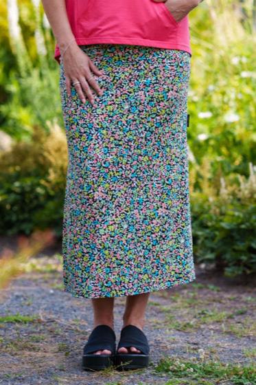 A waist-down view of a woman wearing the Madison 7/8 Skirt by Rien ne se Perd in a Tiny Flowers pattern, featuring a straight cut with side slits and an elastic waist. She's standing outside in front of some plants. 