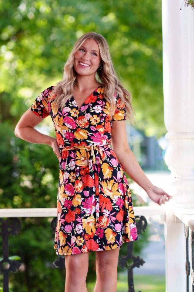 A woman wearing the Margarita Dress by Rien ne se Perd in Red/Orange floral print, with a faux wrap neckline, short sleeves, and short a-line skirt. She is standing on a porch. 