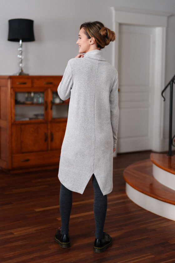 Back view of a woman wearing the Michelangelo Dress by Rien ne se Perd in Pale Grey, a long sleeved turtleneck dress with a heart applique on the front, standing in front of a staircase. 