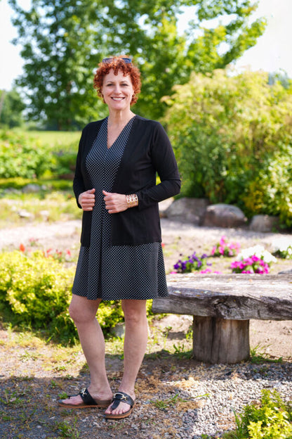 A woman wearing the Samba Dress by Rien ne se Perd in Black and White Polka Dot, a sleeveless short dress with a V-neck, centre pleat, and side pockets. She is wearing it with a black bolero and is standing outside in a tropical setting.