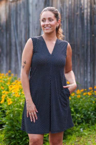 A 3/4 view of a woman wearing the Samba Dress by Rien ne se Perd in Black and White Polka Dot, a sleeveless short dress with a V-neck, centre pleat, and side pockets. She is standing outside in front of a wooden building. 