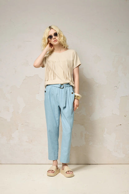 A woman wearing the Samsara Top by Cokluch in Sand, a short sleeved top with fringed V-panel at the chest and a relaxed fit. She is wearing it with blue pants and standing against a neutral background