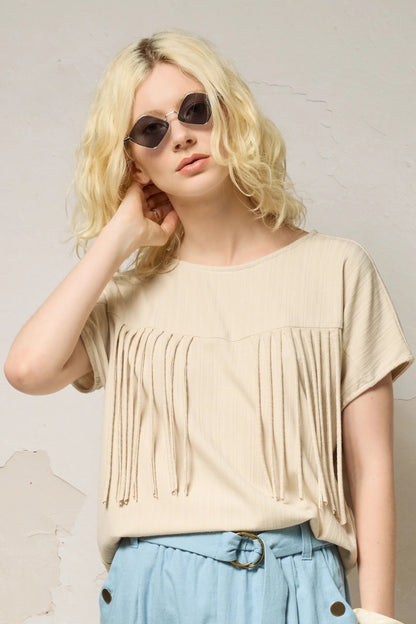 A woman wearing the Samsara Top by Cokluch in Sand, a short sleeved top with fringed V-panel at the chest and a relaxed fit. She is wearing it with blue pants and standing against a neutral background