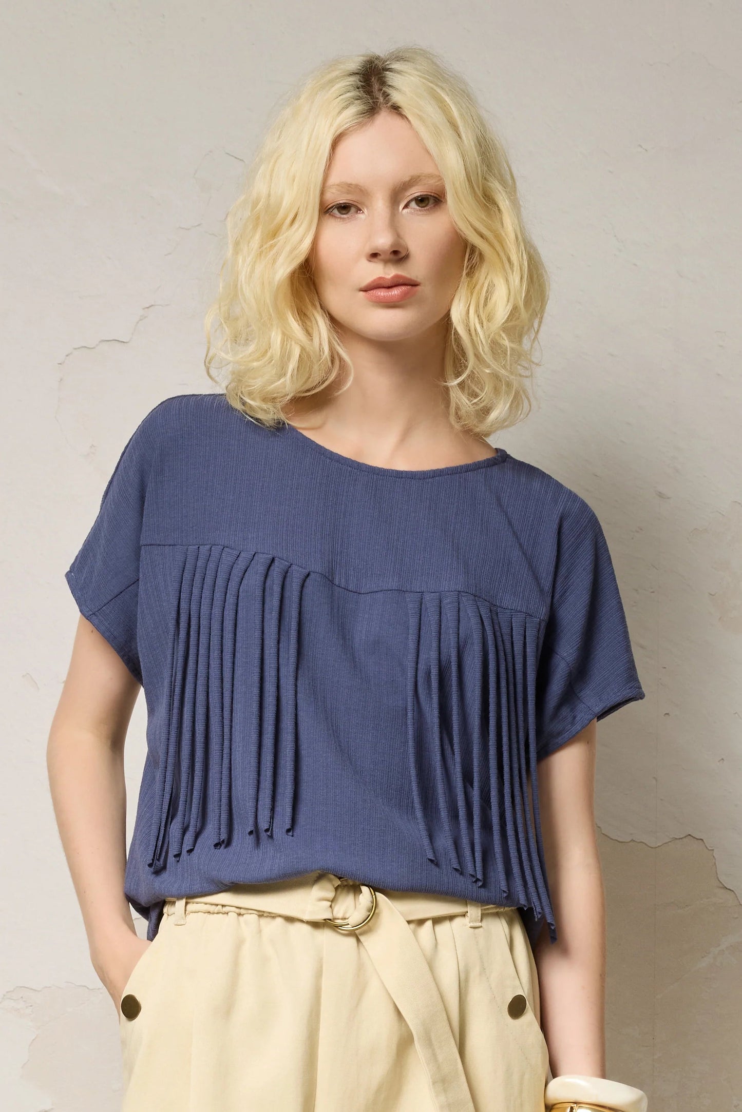 Waist up view of a woman wearing the Samsara Top by Cokluch in Navy, a short sleeved top with fringed V-panel at the chest and a relaxed fit. She is wearing it with cream coloured pants and standing against a neutral background