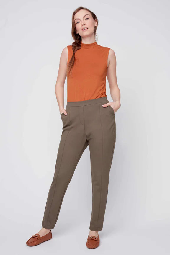 A woman wearing the Sarah Cigarette Pant by Julei in Fern featuring four way stretch fabric, pin tucks for shape, and a pull-on waist. She is wearing them with an orange shirt and standing in front of a white background. 