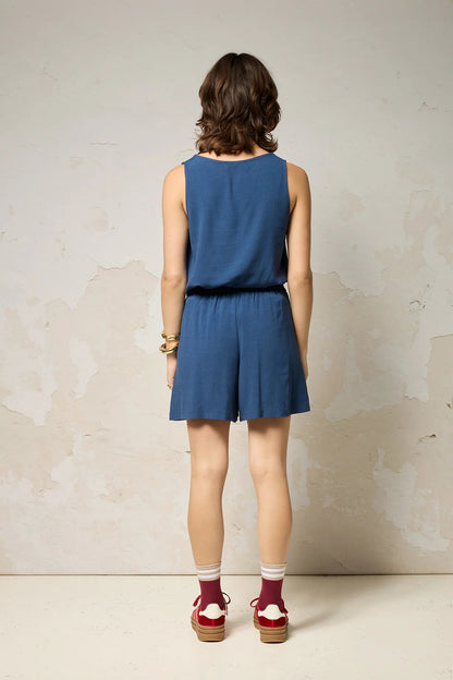 Back view of a woman wearing the Sita Top by Cokluch in Denim, a sleeveless V-neck tank with a loose fit and wide straps. She is wearing it with matching shorts against a plain background