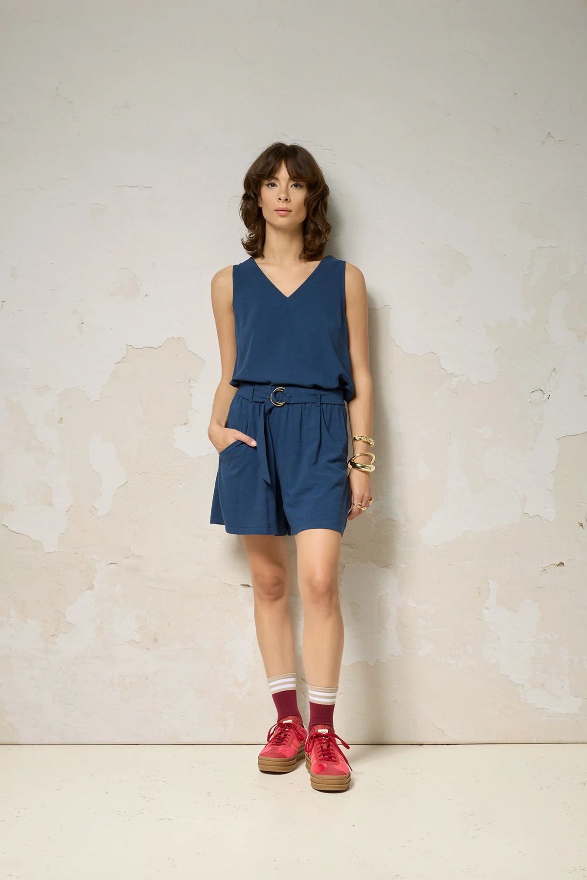 A woman wearing the Atman Shorts by Cokluch in Denim blue, mid-thigh length shorts with a high elastic waist and an attached ring belt. She is wearing them with a matching top and standing in front of a plain background. 