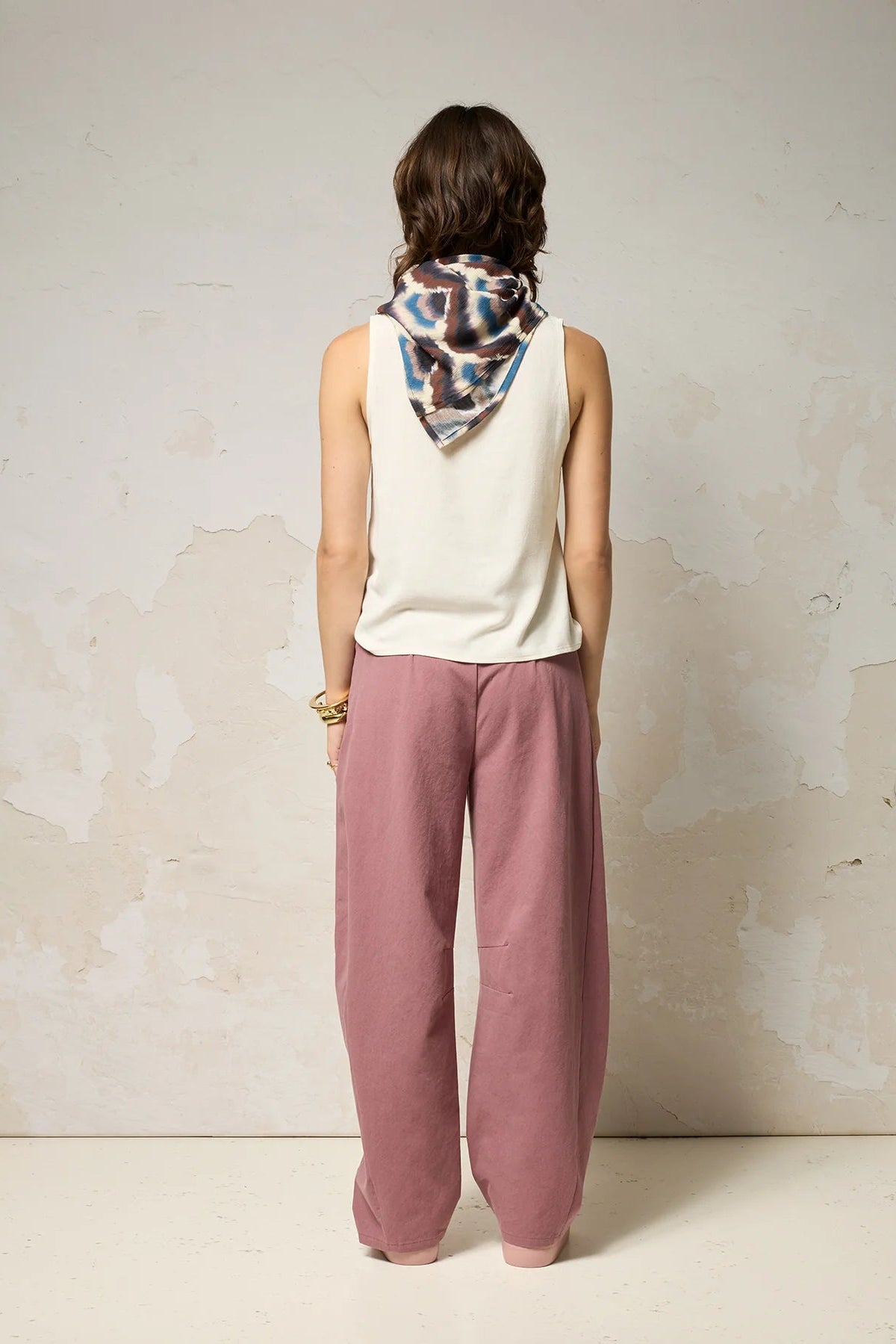 Back view of a woman wearing the Sita Top by Cokluch in Natural a sleeveless V-neck tank with a loose fit and wide straps. She is wearing it with pink pants and a patterned scarf against a plain background