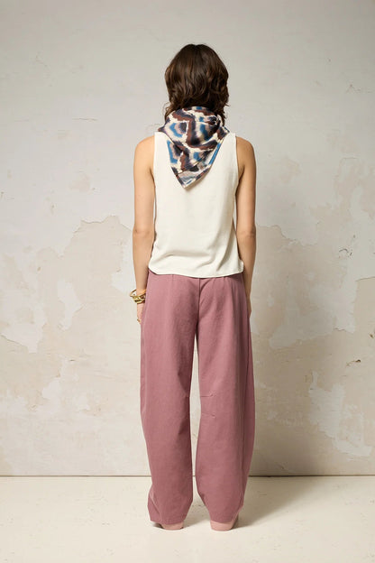Back view of a woman wearing the Sita Top by Cokluch in Natural a sleeveless V-neck tank with a loose fit and wide straps. She is wearing it with pink pants and a patterned scarf against a plain background