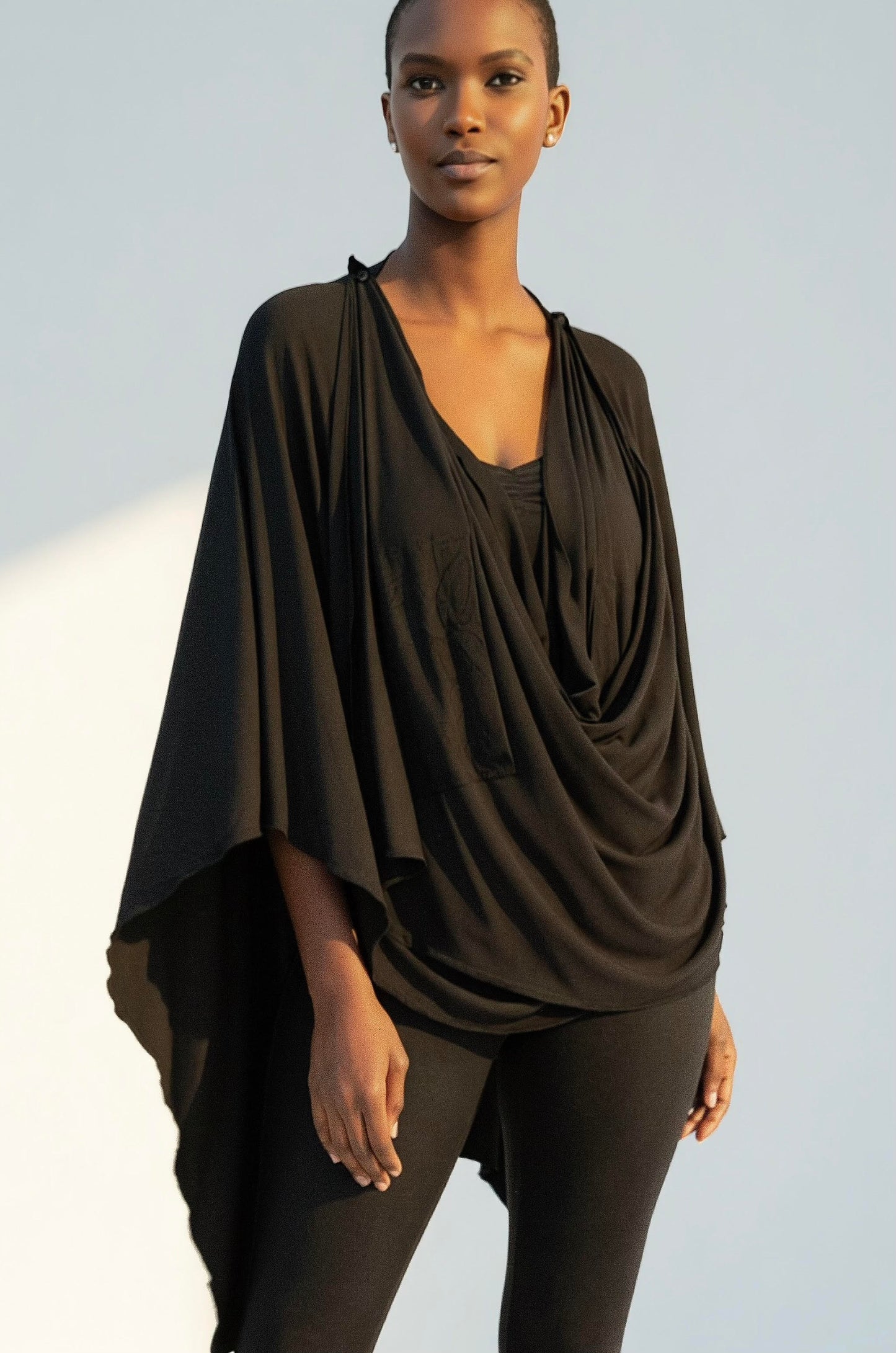 A woman wearing the Endless Bamboo Multi-wrap top by Stacey Martin Lifestyle in Black, a top that can be wrapped and fastened by the shoulder buttons in many different ways. She is wearing it with black leggings and standing against a grey background. 