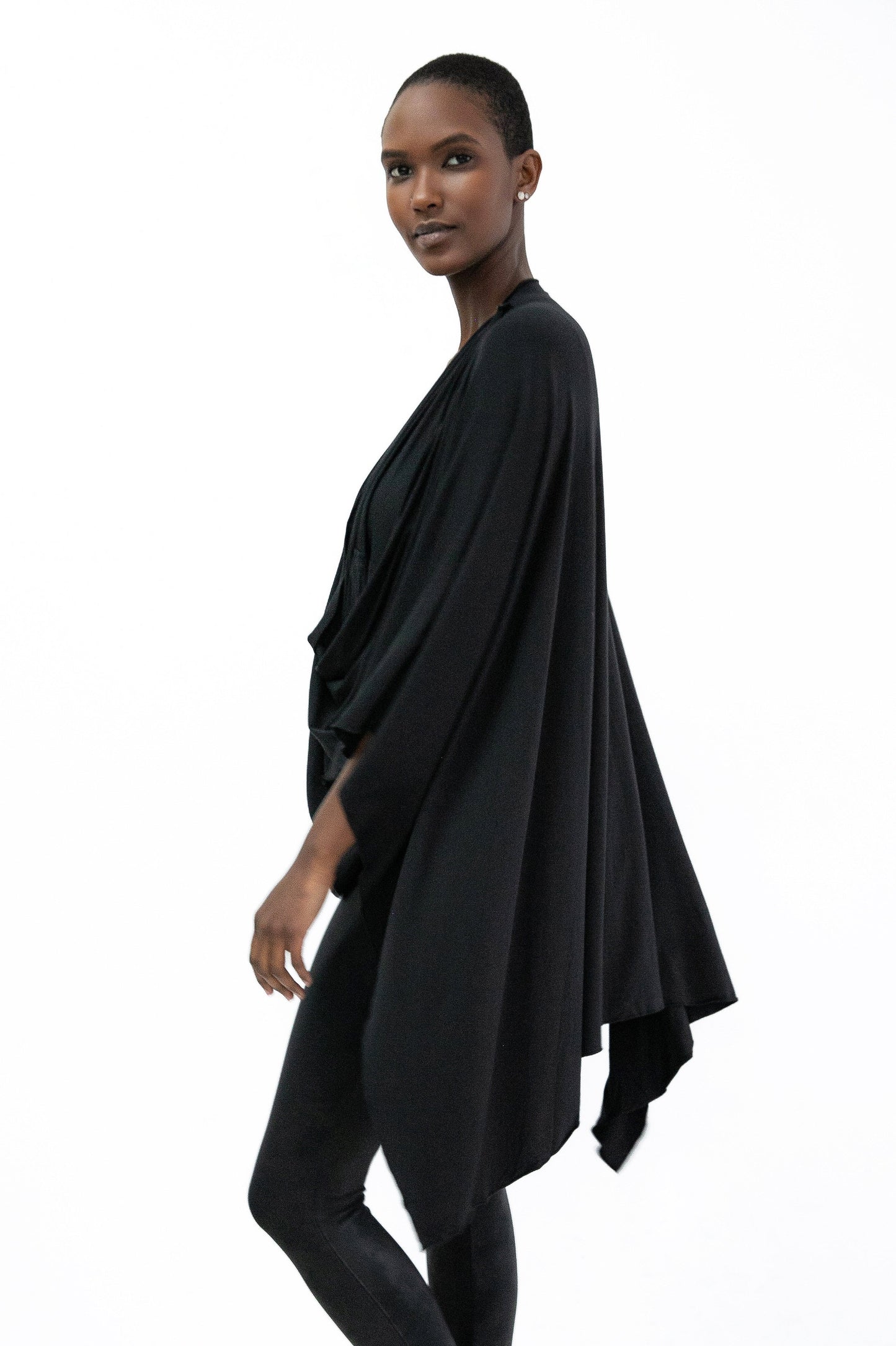 Side view of a woman wearing the Endless Bamboo Multi-wrap top by Stacey Martin Lifestyle in Black, a top that can be wrapped and fastened by the shoulder buttons in many different ways. She is wearing it with black leggings and standing against a white background. 