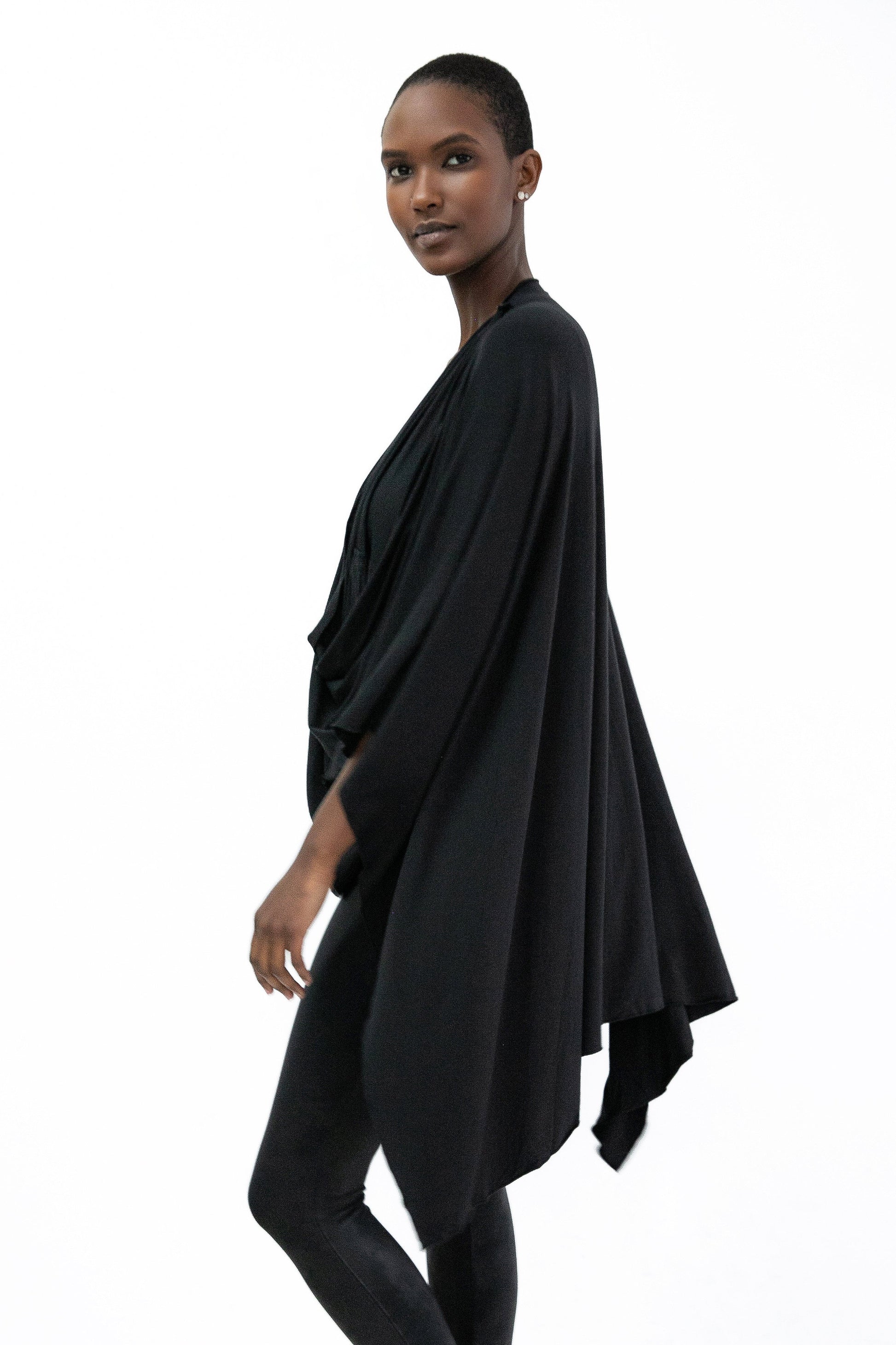 Side view of a woman wearing the Endless Bamboo Multi-wrap top by Stacey Martin Lifestyle in Black, a top that can be wrapped and fastened by the shoulder buttons in many different ways. She is wearing it with black leggings and standing against a white background. 