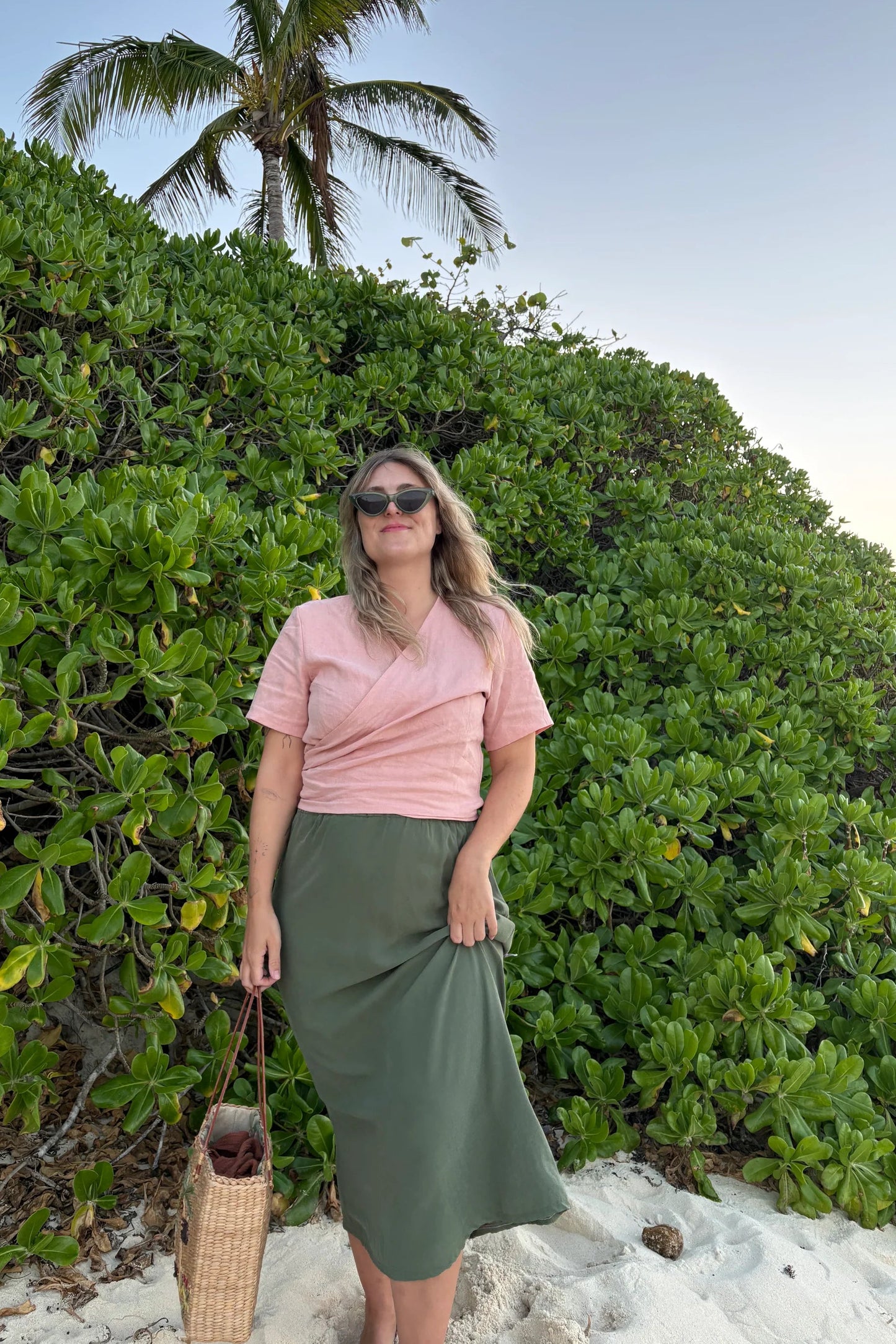 A woman wearing the Simone Skirt by Lights of All in Cactus, a sleek calf-length skirt with a flared hem and an elastic waist. She is wearing it with the Stella Top in Ballet Pink and standing on a beach in front of foliage.