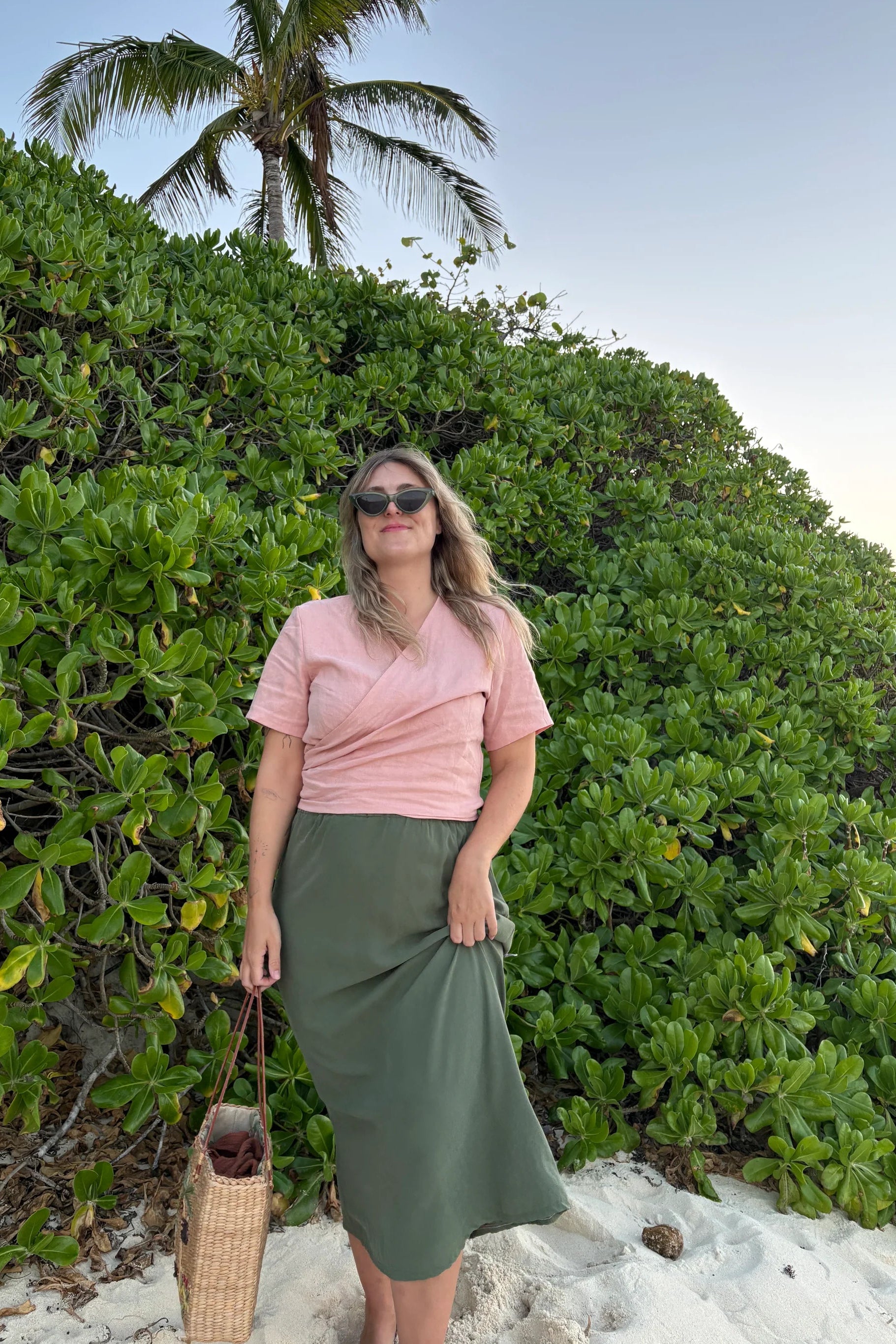 A woman wearing the Simone Skirt by Lights of All in Cactus, a sleek calf-length skirt with a flared hem and an elastic waist. She is wearing it with the Stella Top in Ballet Pink and standing on a beach in front of foliage.