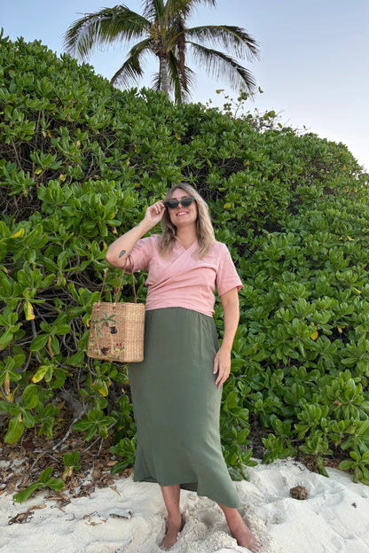 A woman wearing the Simone Skirt by Lights of All in Cactus, a sleek calf-length skirt with a flared hem and an elastic waist. She is wearing it with the Stella Top in Ballet Pink and standing on a beach in front of foliage.